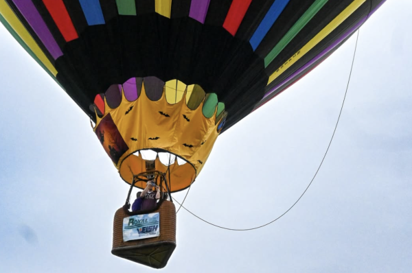 A colorful hot air balloon with a striped pattern floats in the sky during a Hot Air Balloon Festival. Two people are in the basket with a Royal LePage sign, viewed from below against a clear blue background.